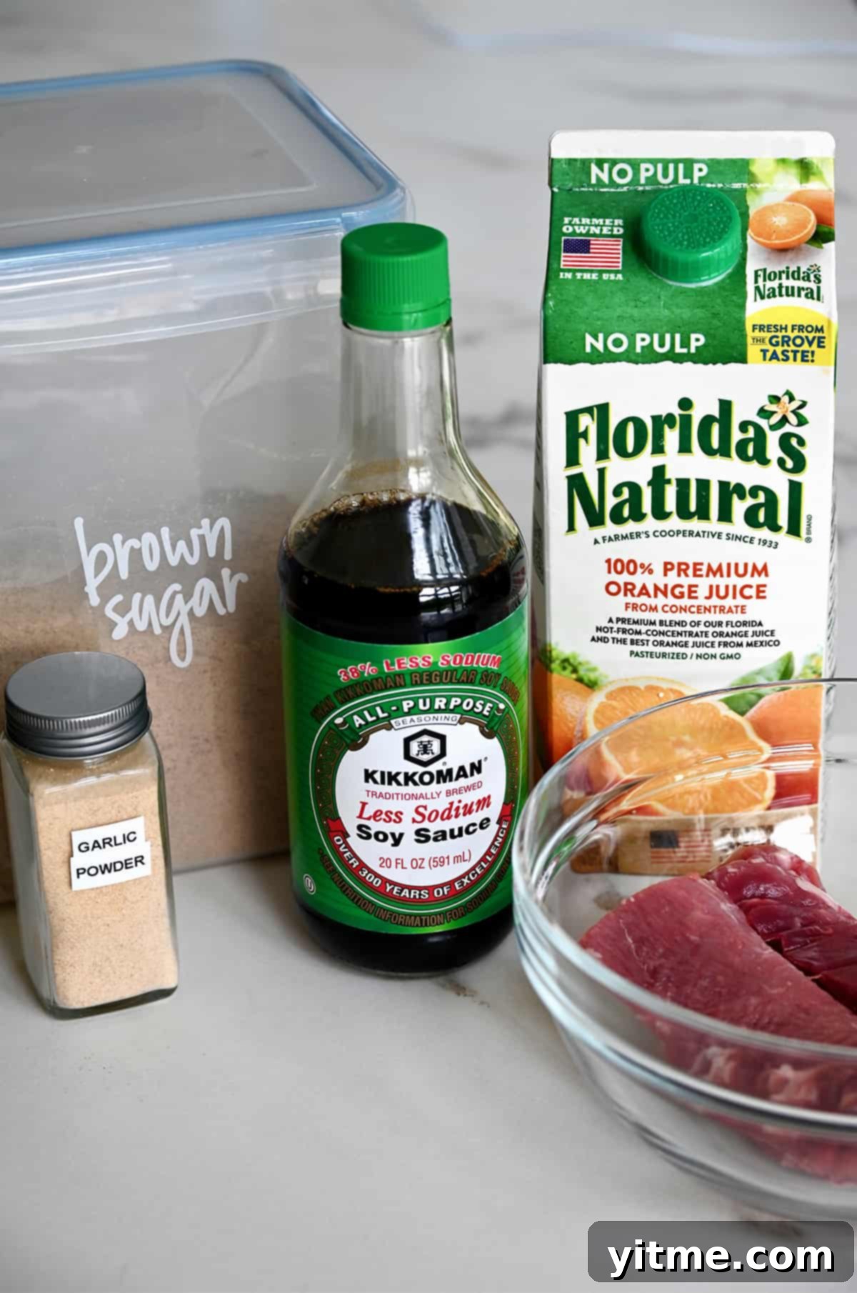 A small jar containing garlic powder next to a plastic container with brown sugar, a bottle of low sodium soy sauce, a carton of Florida's Natural orange juice and a clear bowl with raw pork tenderloin.