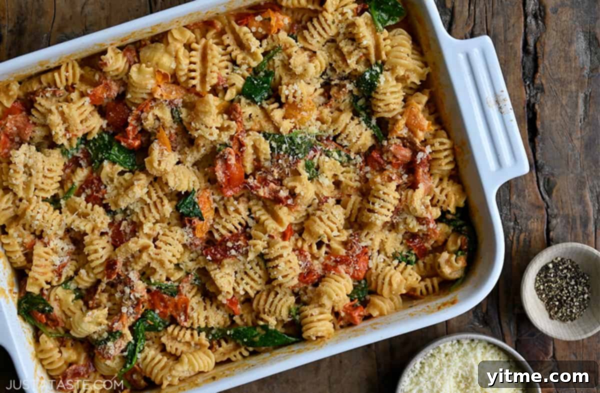 A top-down view of Baked Boursin Pasta with roasted tomatoes and spinach in a white baking dish next to small bowls containing black pepper and Parmesan cheese.