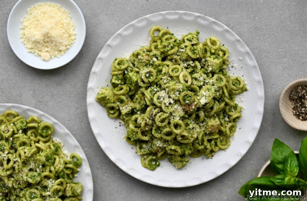 A white dinner plate containing a generous serving of pesto pasta with chicken, artfully arranged next to a small dish of grated Parmesan cheese for topping.