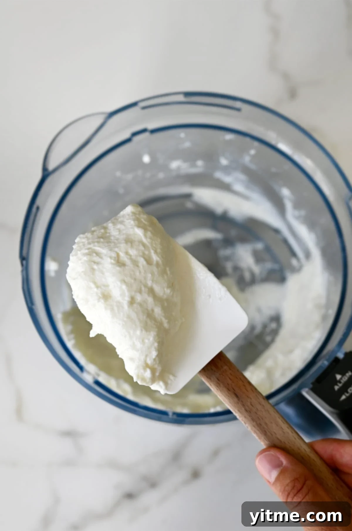 Whipped cottage cheese on a spatula above a food processor.