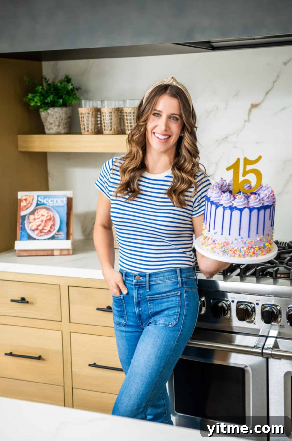 Chef Kelly Senyei standing in her kitchen holding a purple cake with the number "15" on it, celebrating Just a Taste\
