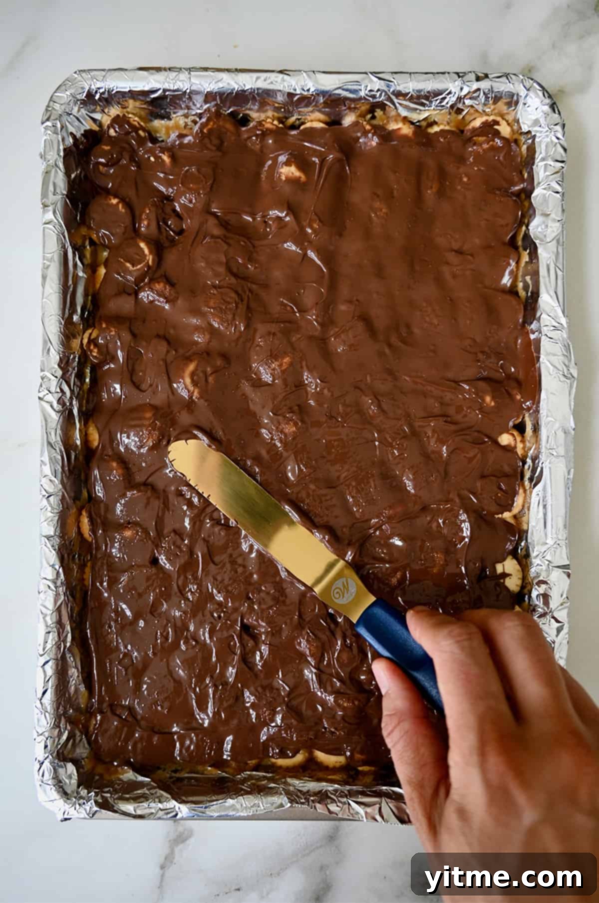 A hand holds an offset spatula to spread melted chocolate atop toffee-coated oyster crackers on a foil-lined baking sheet.