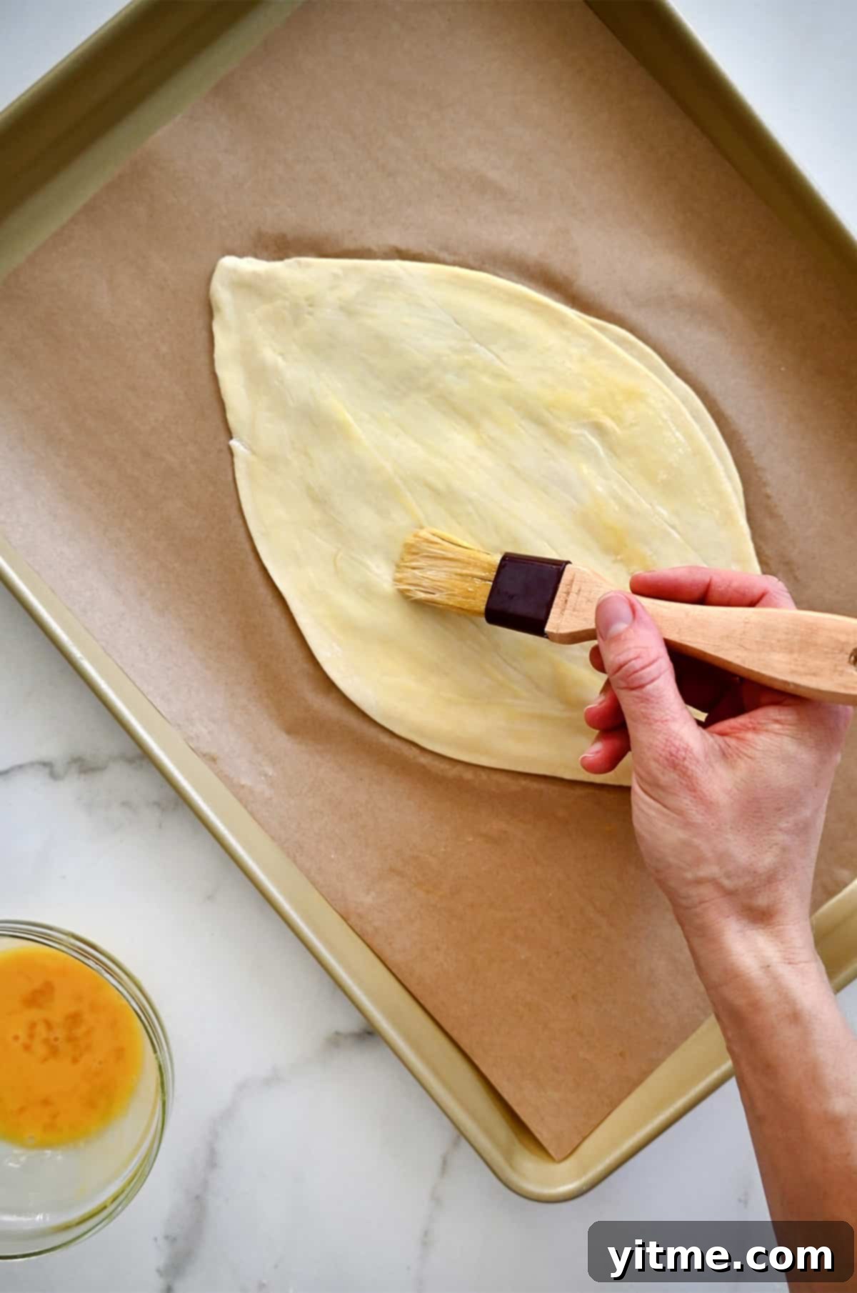 A hand holding a pastry brush applies an egg wash to football-shaped puff pastry on a parchment paper-lined baking sheet.