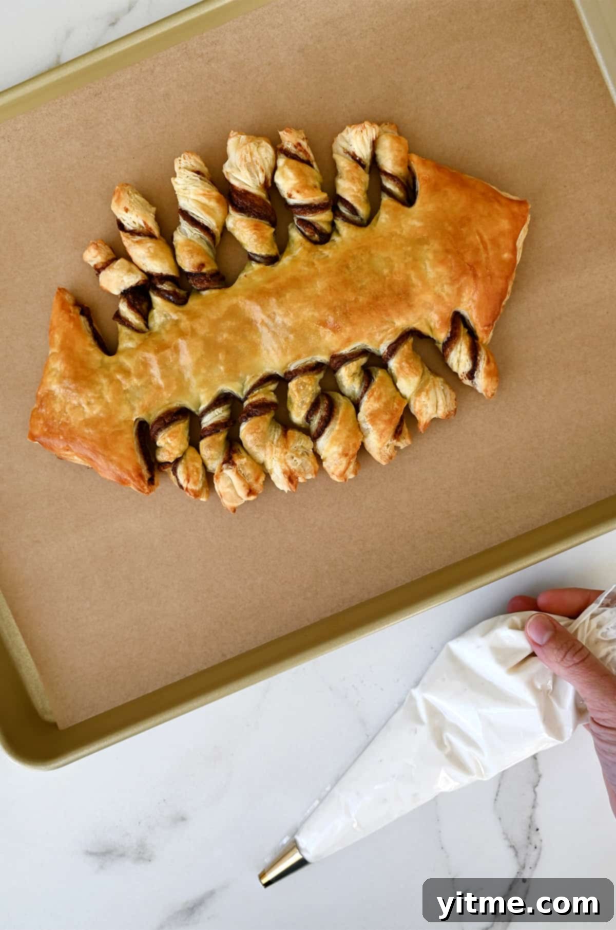 A football-shaped puff pastry with Nutella filling on a parchment paper-lined baking sheet next to a piping bag with vanilla frosting.