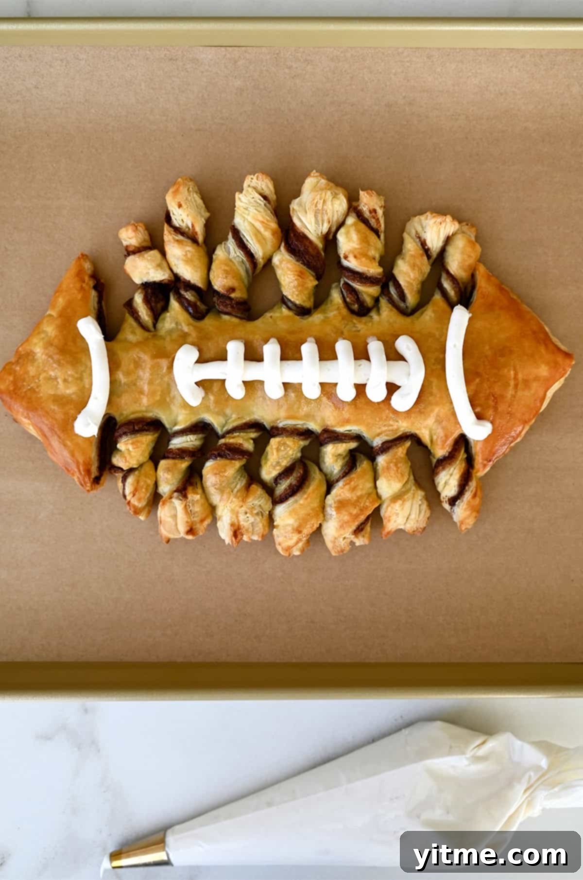 Pastry football with vanilla frosting laces on a baking sheet next to a piping bag filled with frosting.