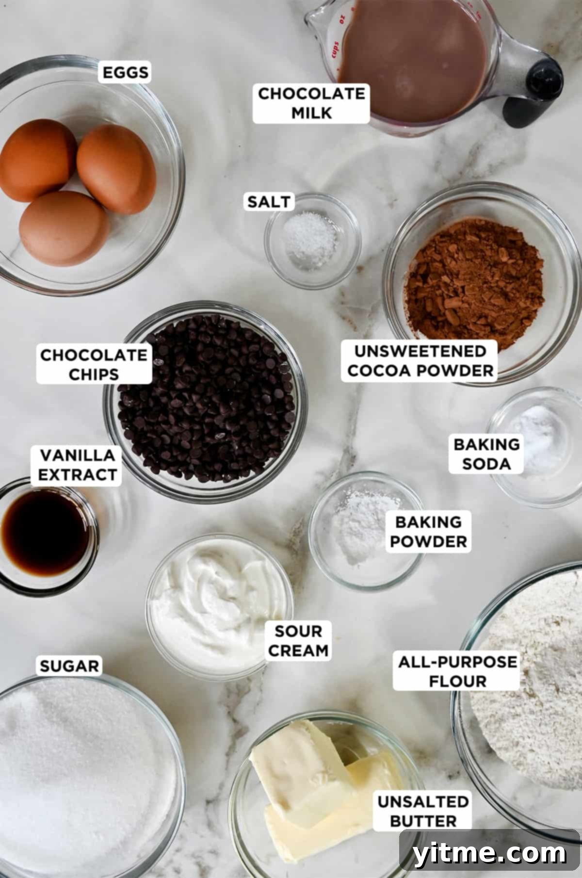 Various sizes of glass bowls containing the ingredients needed to make chocolate cupcakes, including three eggs, chocolate milk, salt, unsweetened cocoa powder, baking soda, baking powder, all-purpose flour, sour cream, chocolate chips, vanilla extract, sugar and unsalted butter.