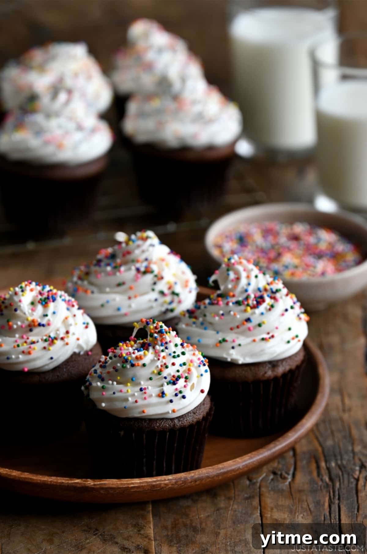 Four chocolate cupcakes topped with white frosting and rainbow sprinkles on a plate with more cupcakes on a wire rack in the background.