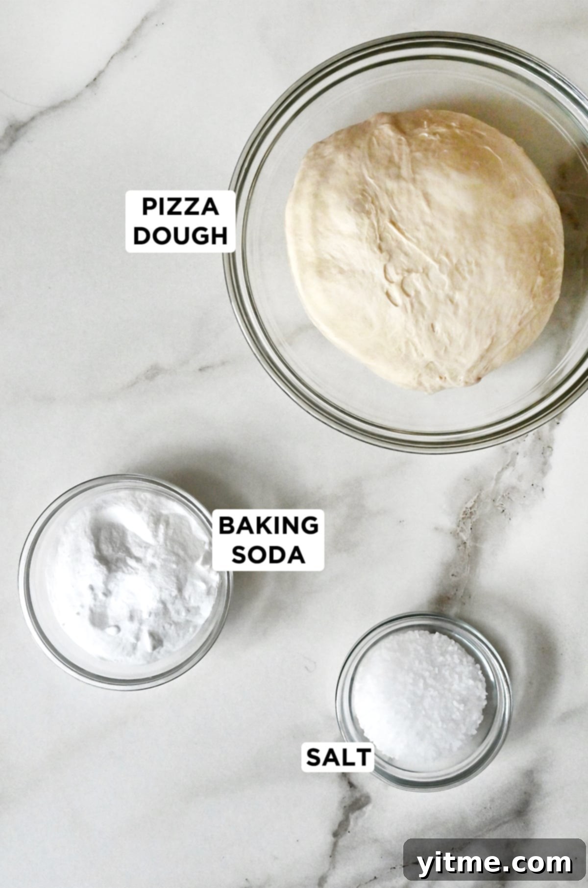 Three glass bowls containing the key ingredients: fresh pizza dough, baking soda, and kosher salt, neatly arranged on a white surface.
