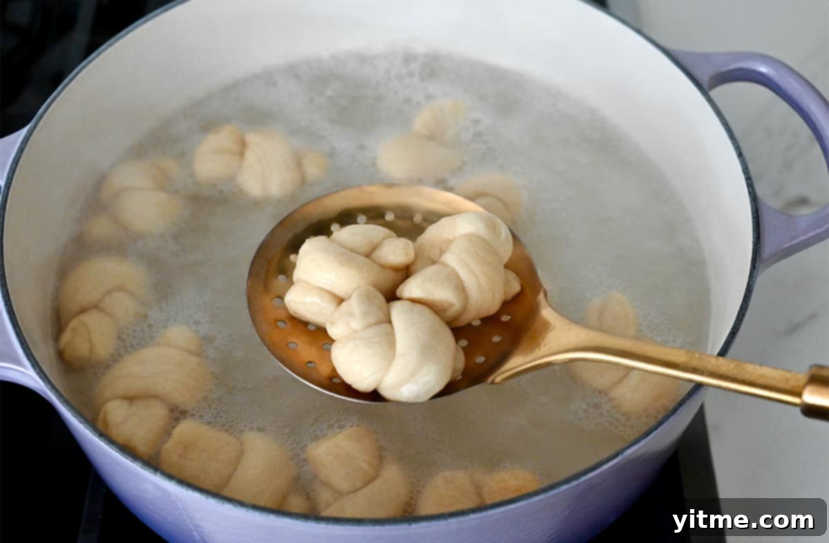 A slotted spoon lifts a boiled pretzel knot from a Dutch oven filled with a bubbling baking soda bath, ready to be placed on a baking sheet.