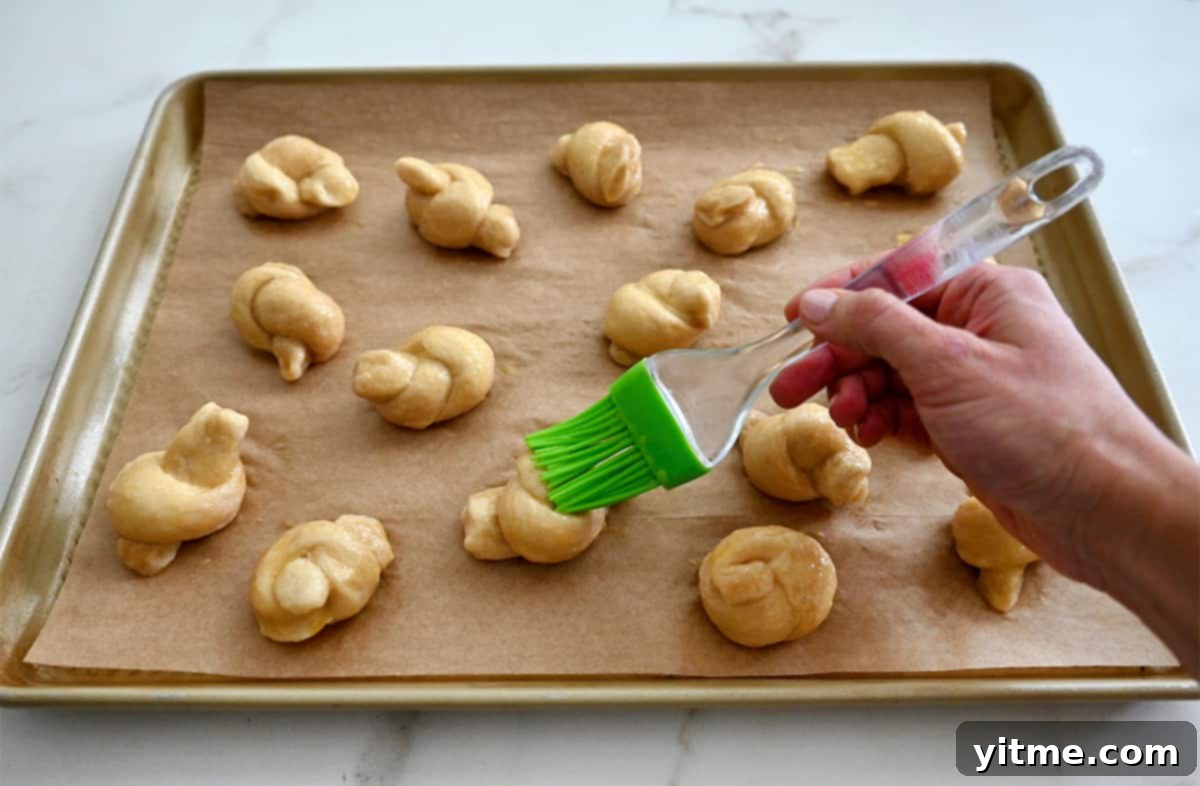 A hand gently brushes an egg wash onto a row of pizza dough knots on a marble surface, preparing them for baking and salting.