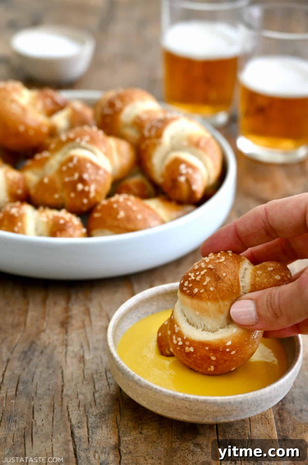 A hand holds a freshly baked pizza dough soft pretzel knot, dipping it into a creamy cheese sauce, with a bowl of other pretzel knots in the blurred background.