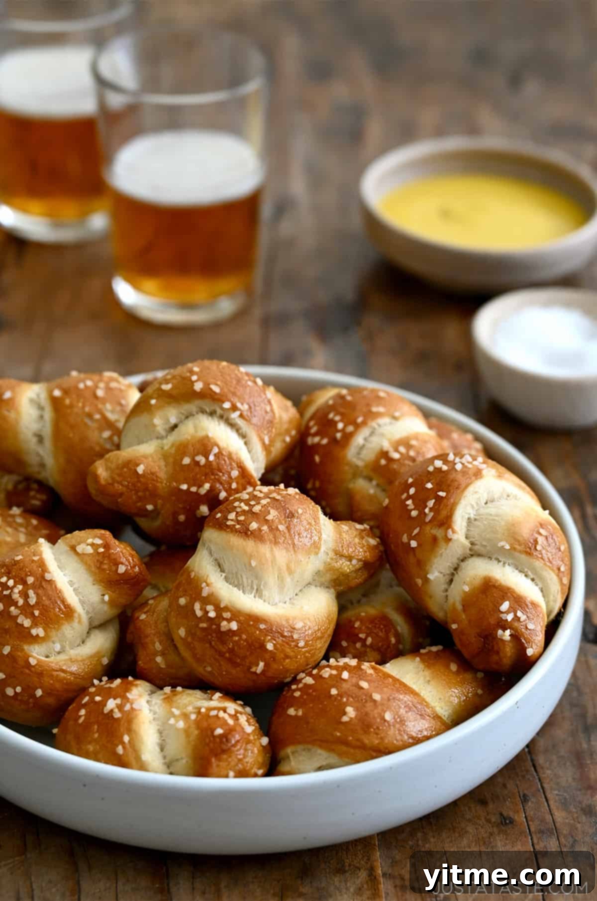 Pizza dough soft pretzel knots piled high in a white bowl with two glasses of beer nearby, perfect for a casual snack or appetizer.
