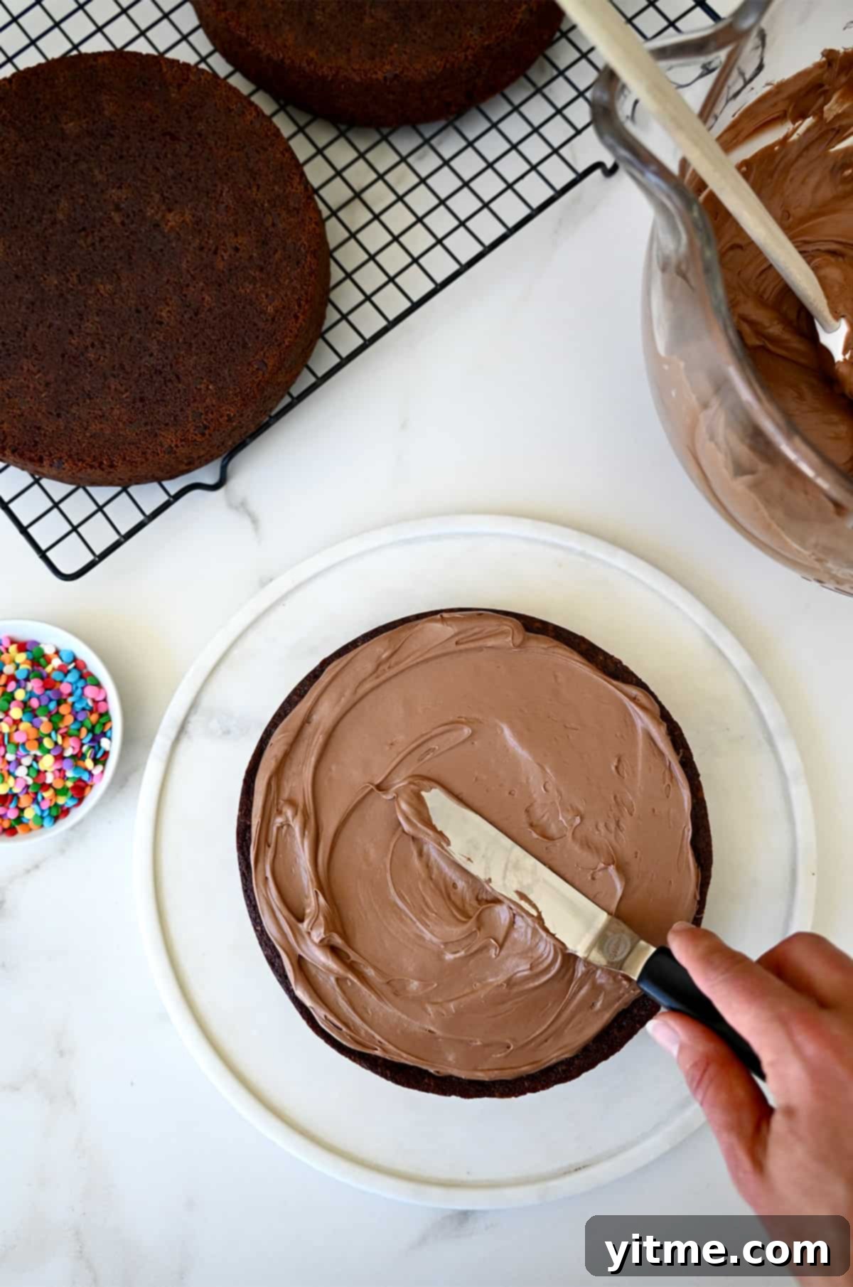 A hand spreading coffee buttercream frosting atop a chocolate cake.