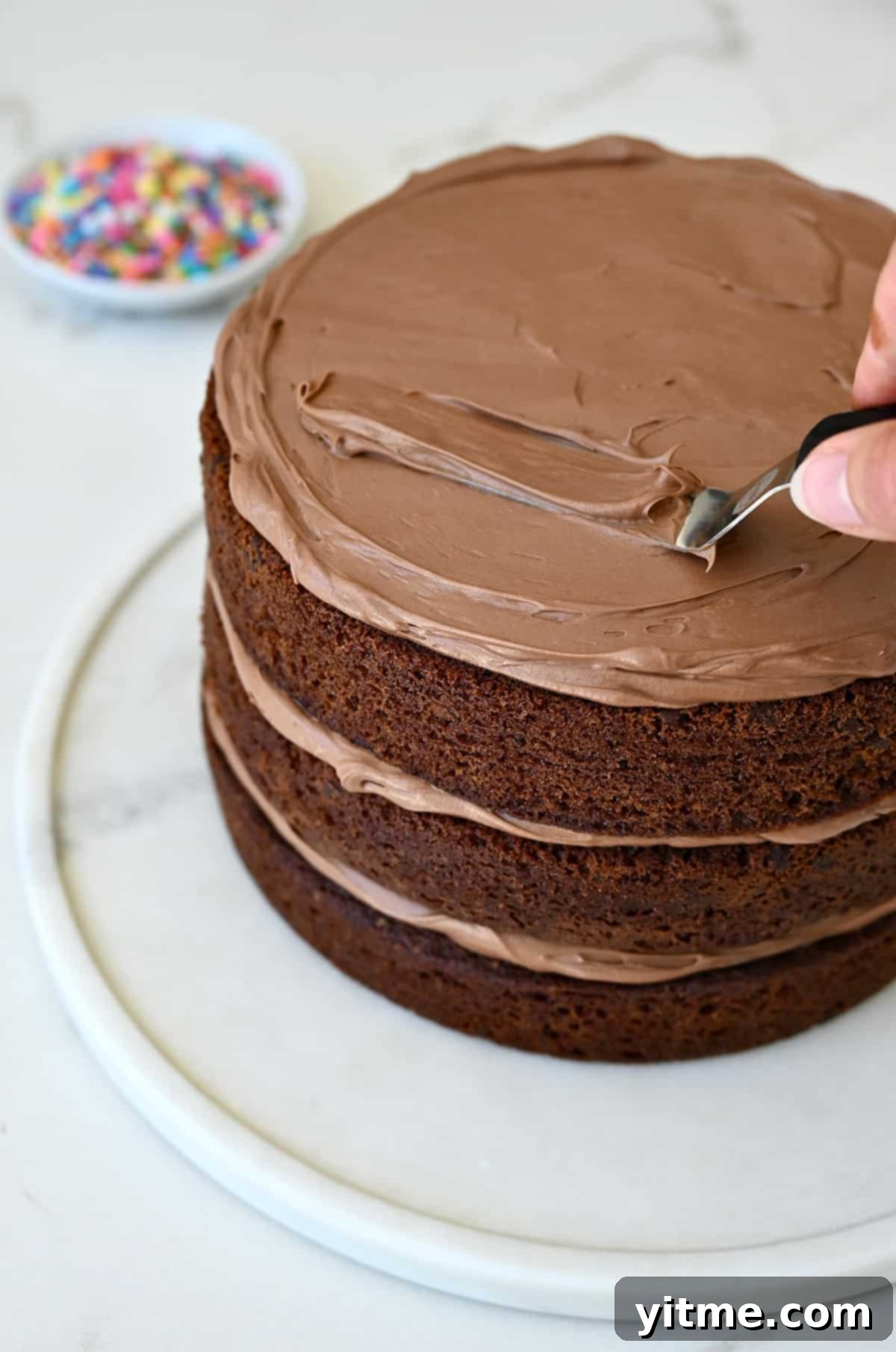 A hand spreading coffee buttercream to the top of a three-layer chocolate cake.