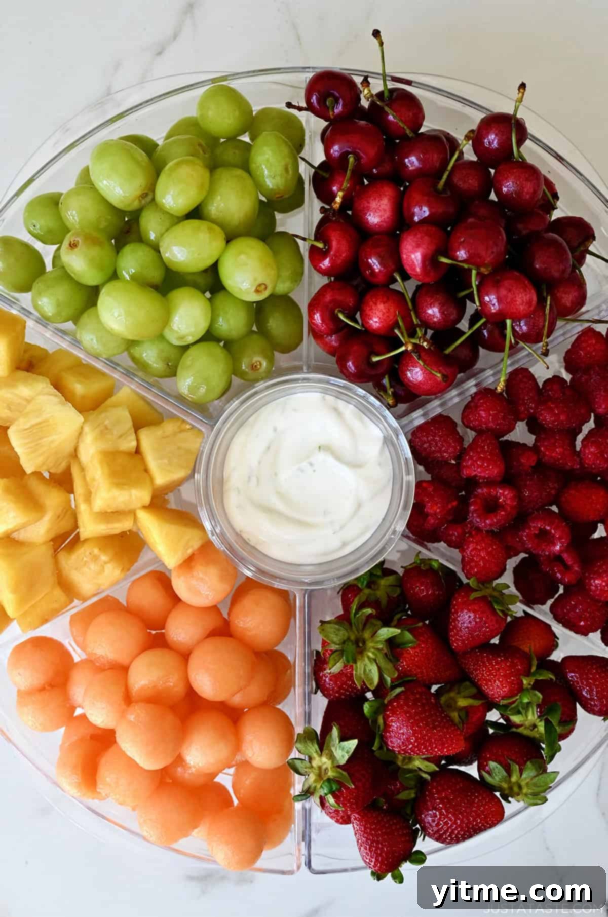 A serving tray with six compartments filled with green grapes, cherries, raspberries, strawberries, cantaloupe balls and chopped pineapple. A small bowl in the center is filled with yogurt.
