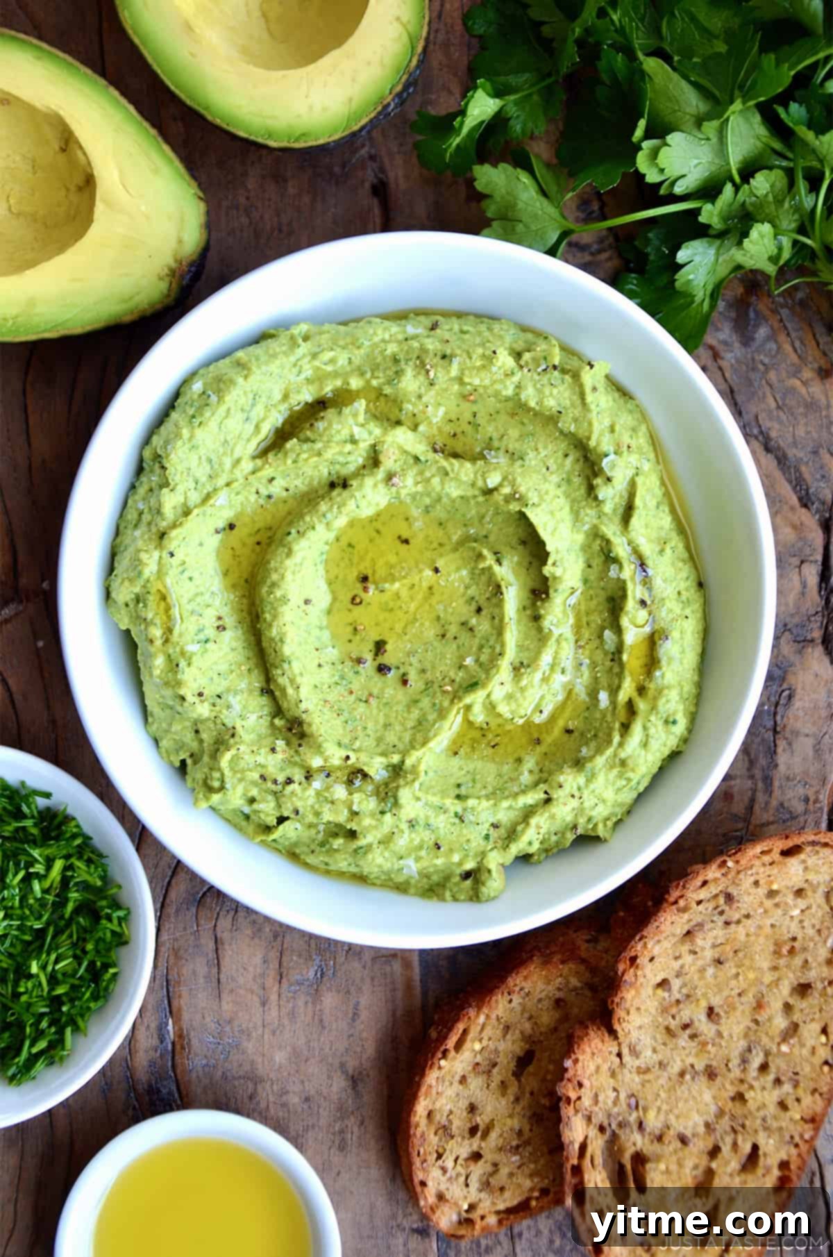 Avocado hummus in a bowl topped with olive oil, flakey salt and pepper. Small bowls of olive oil and minced chives, toasted bread, fresh parsley and a halved avocado surround the hummus.