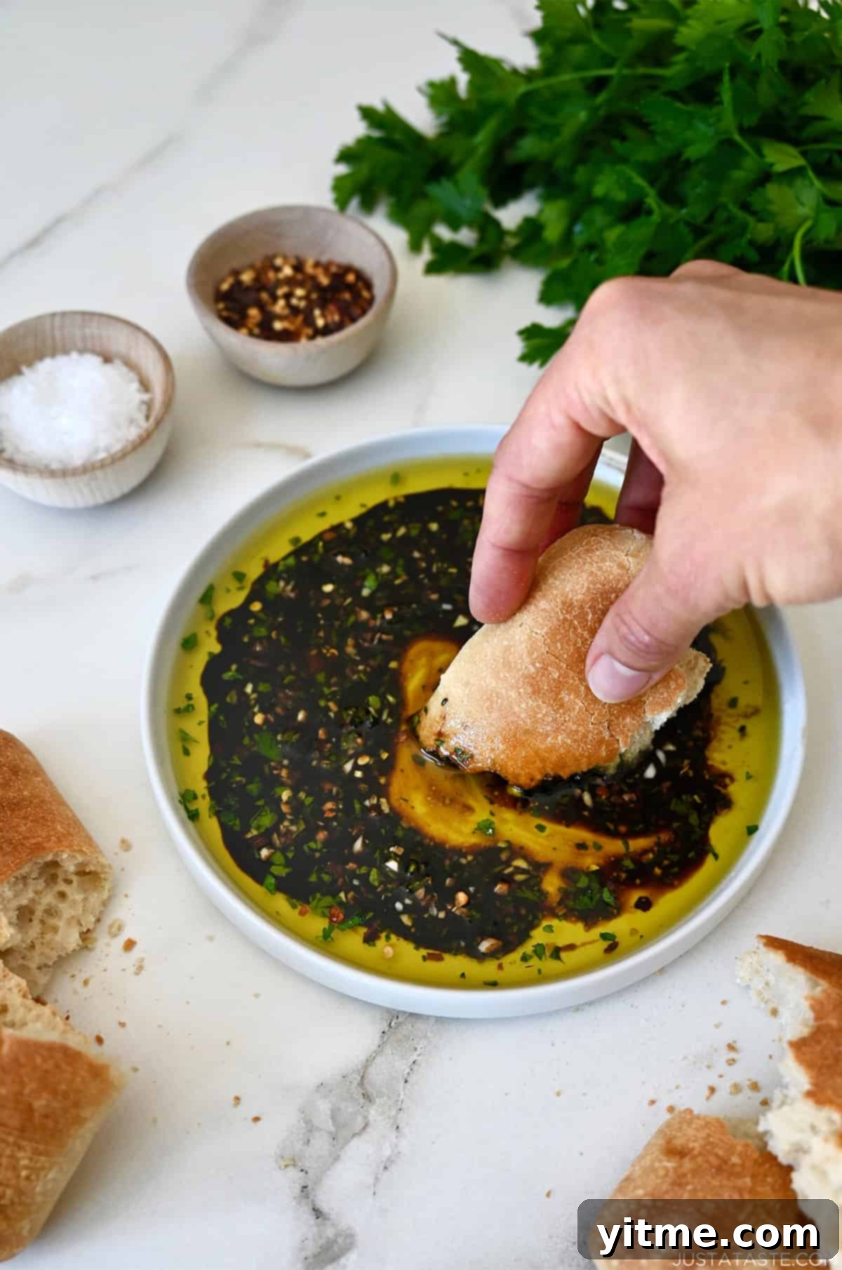 A hand holding a piece of ciabatta bread dips it into a mixture of olive oil, balsamic vinegar and spices on a plate.