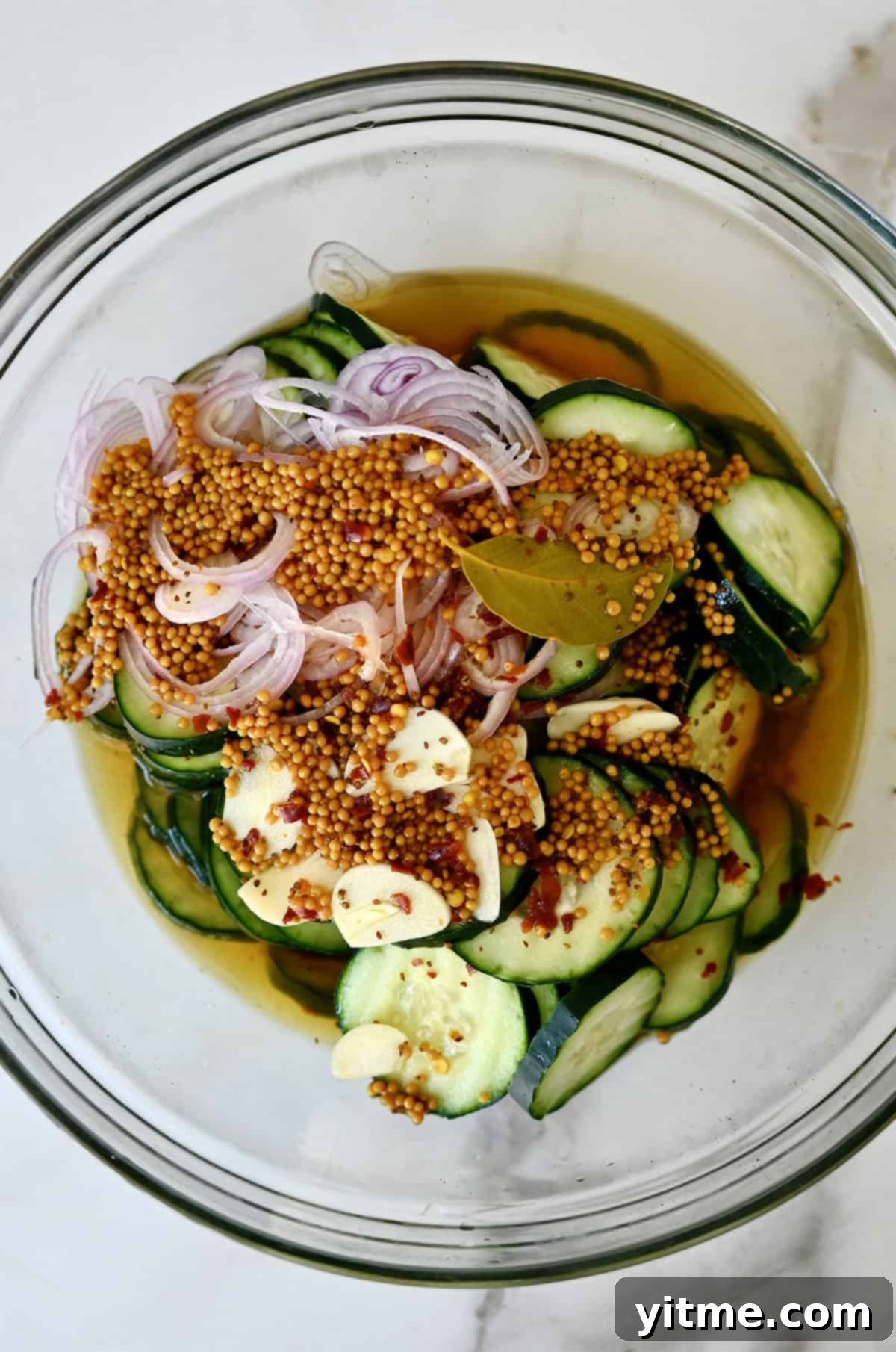 A glass bowl containing thinly sliced zucchini, shallots, and garlic cloves covered in a pickling brine.