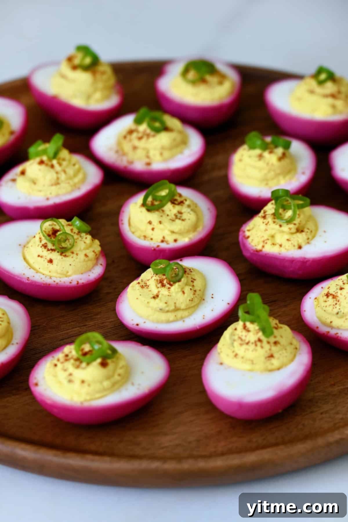 Pickled deviled eggs dyed pink with beets, garnished with paprika and scallions, arranged on a wooden board.