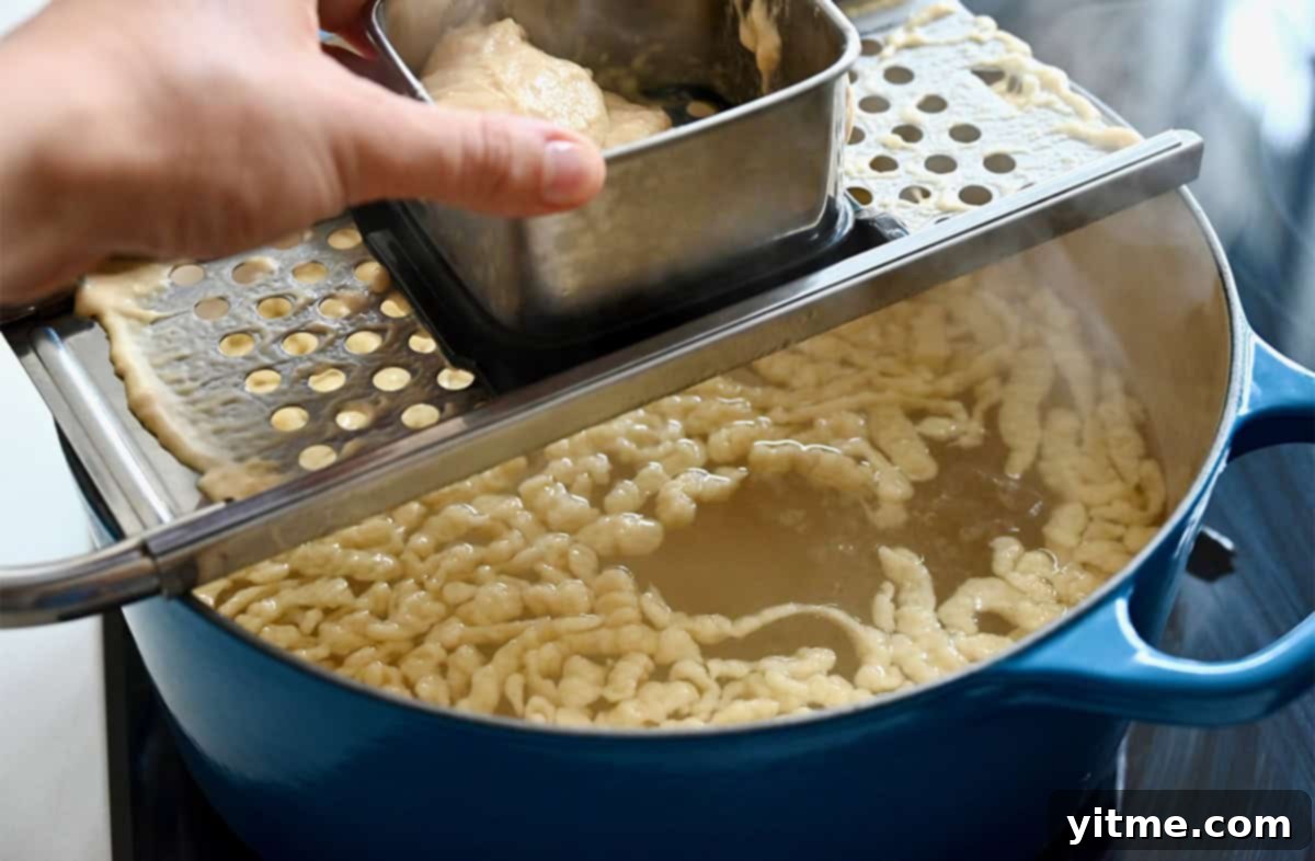 Scraping spaetzle dough over a spaetzle maker that sits on top of a large pot with boiling water.