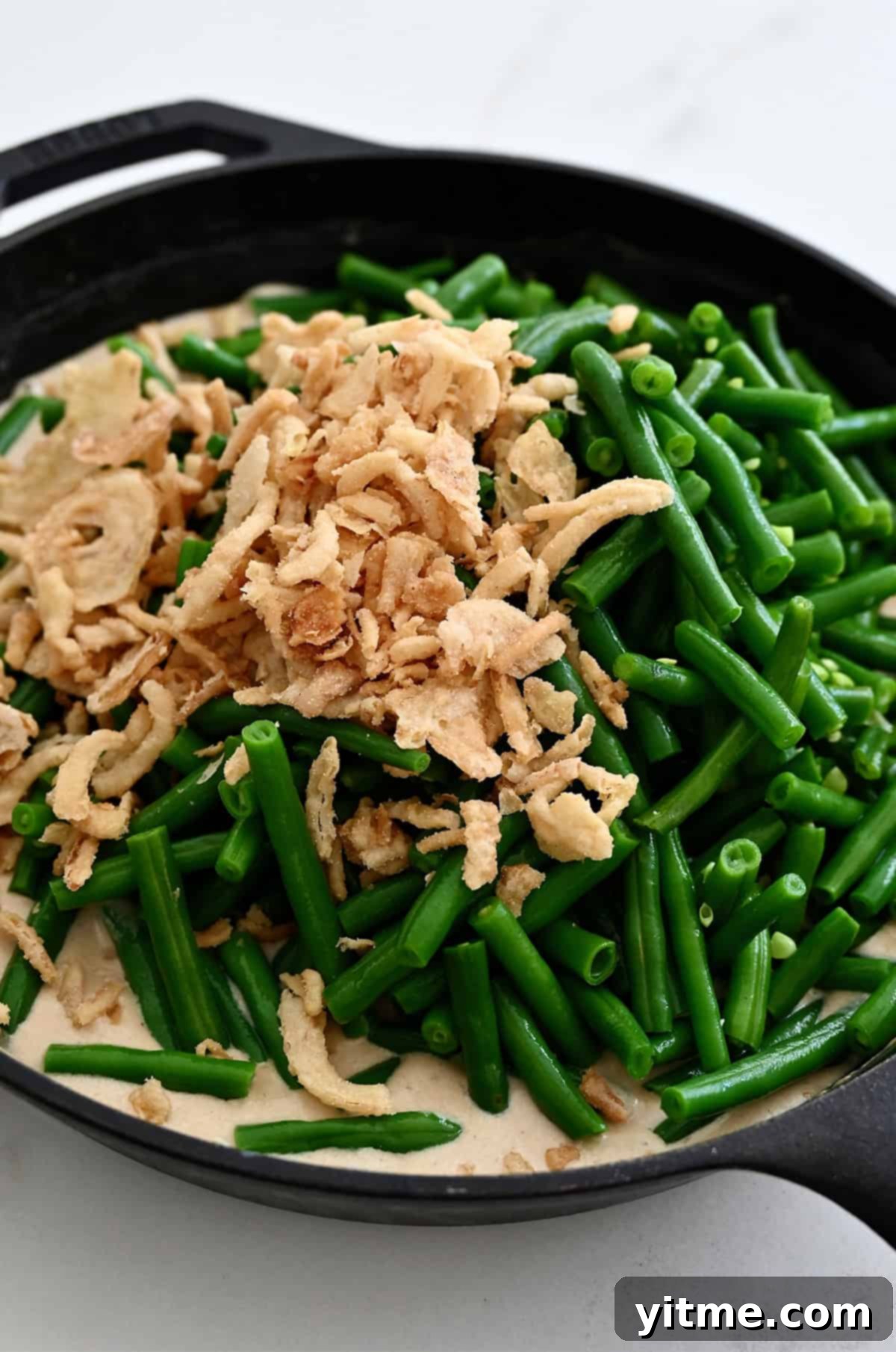 Green beans and crispy fried onions mixed into homemade cream of mushroom soup in a skillet, ready for baking.