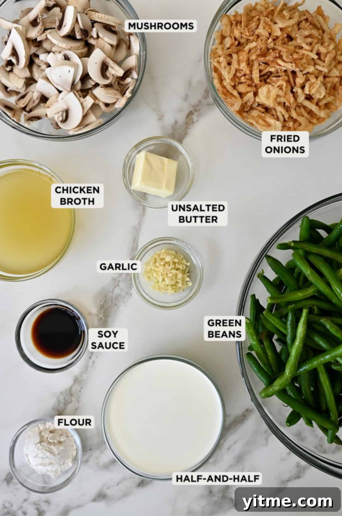 Green bean casserole ingredients in glass bowls, including fresh green beans, flour, half-and-half, mushrooms, butter, chicken broth, soy sauce and crispy fried onions.