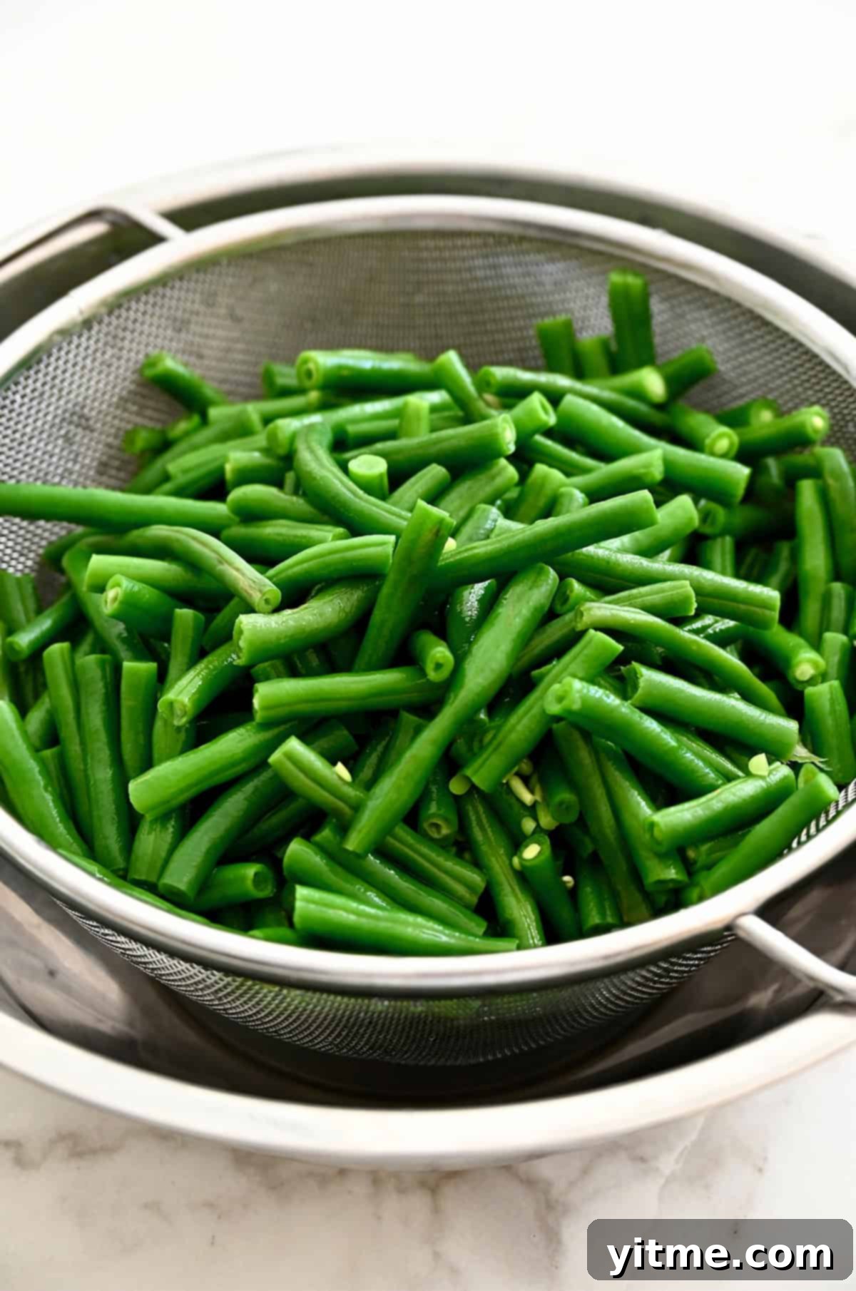 Blanched green beans draining thoroughly in a sieve over a bowl.