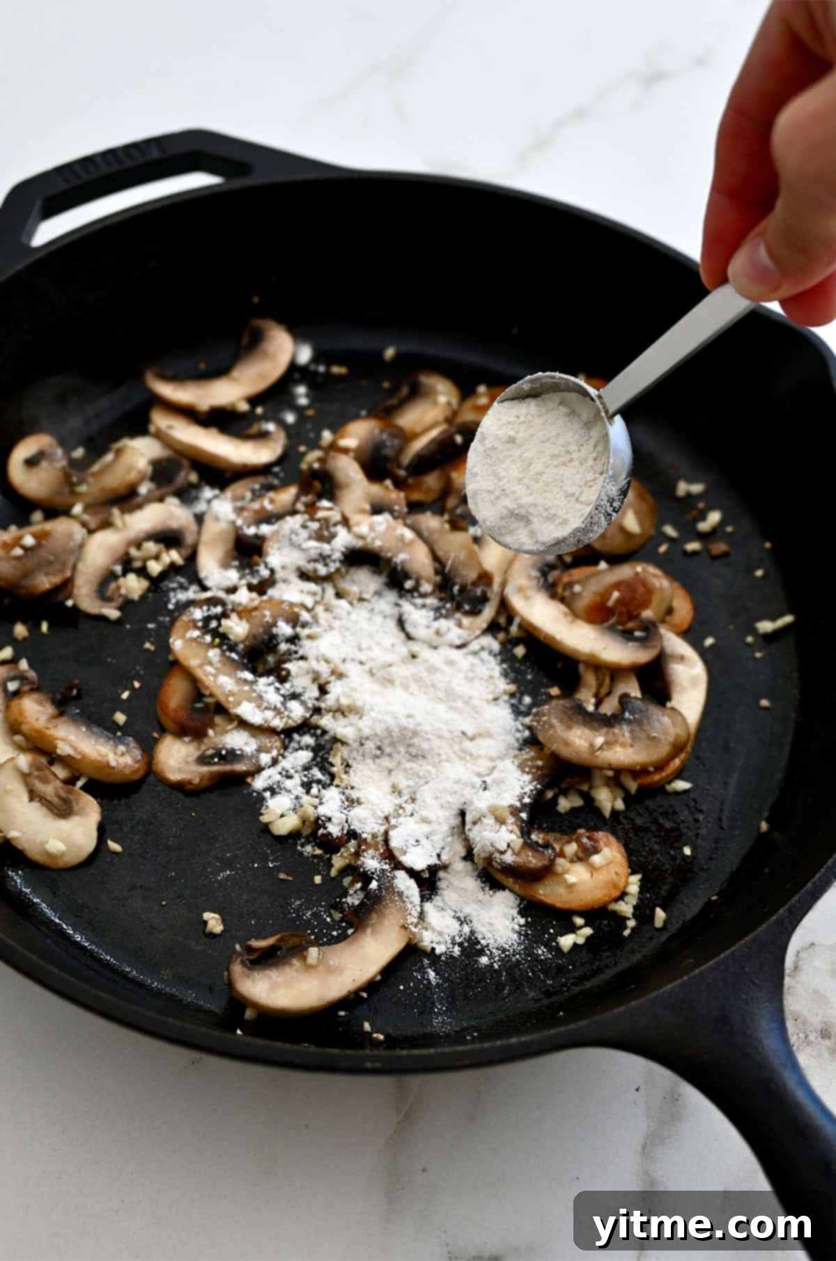 Flour being stirred into sautéed mushrooms and garlic in a skillet to form a roux.