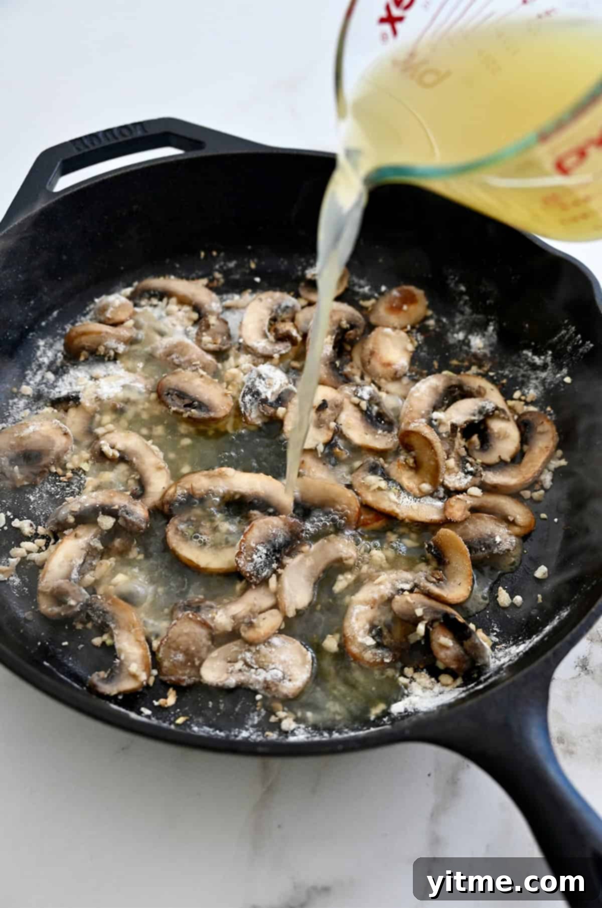Chicken broth being poured into a skillet with mushroom-flour mixture for sauce.