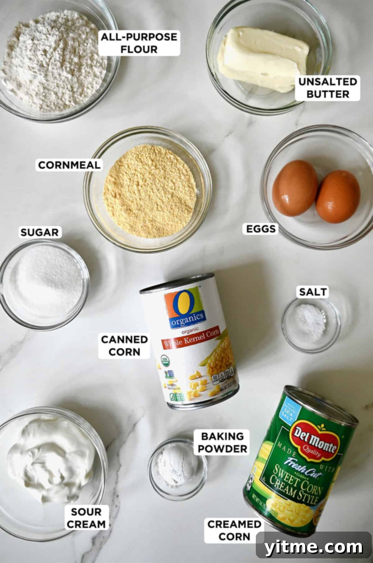 Various ingredients for creamy corn casserole, including bowls of flour, cornmeal, sugar, sour cream, and eggs, alongside cans of creamed corn and whole kernel corn.