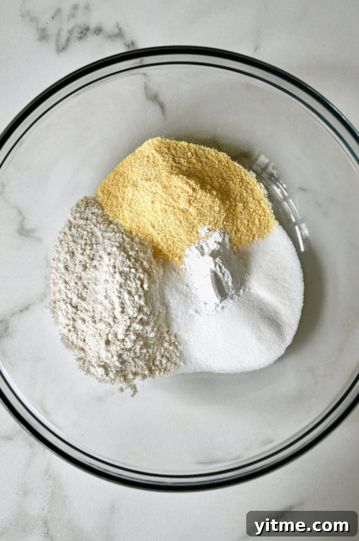 A glass bowl containing dry ingredients for corn casserole: yellow cornmeal, all-purpose flour, baking powder, sugar, and a pinch of salt.