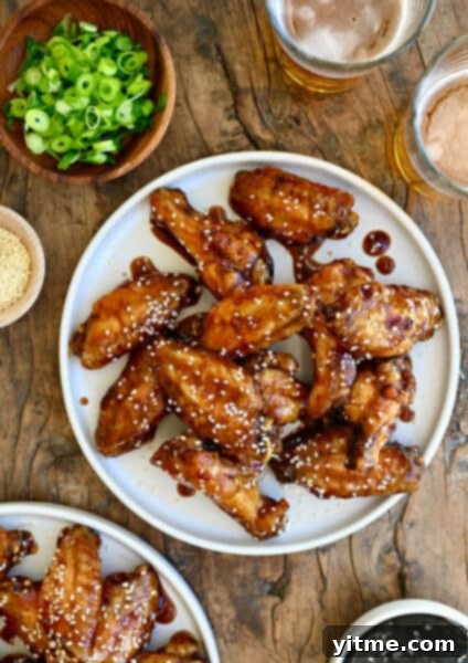 Teriyaki chicken wings garnished with sesame seeds on a white plate next to two glasses containing beer and a small bowl with sliced scallions.