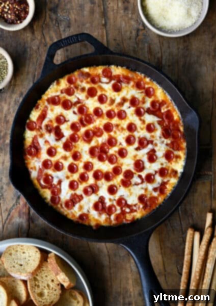 Pepperoni pizza dip in a cast iron skillet next to a small bowl containing crispy baguette toasts.