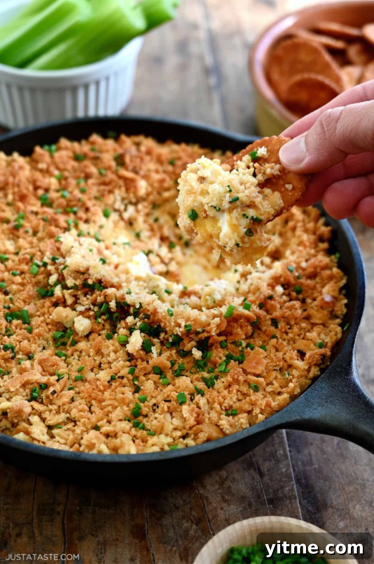 A hand holding a Ritz cracker dips it into a cheesy jalapeño popper dip in a cast iron skillet.