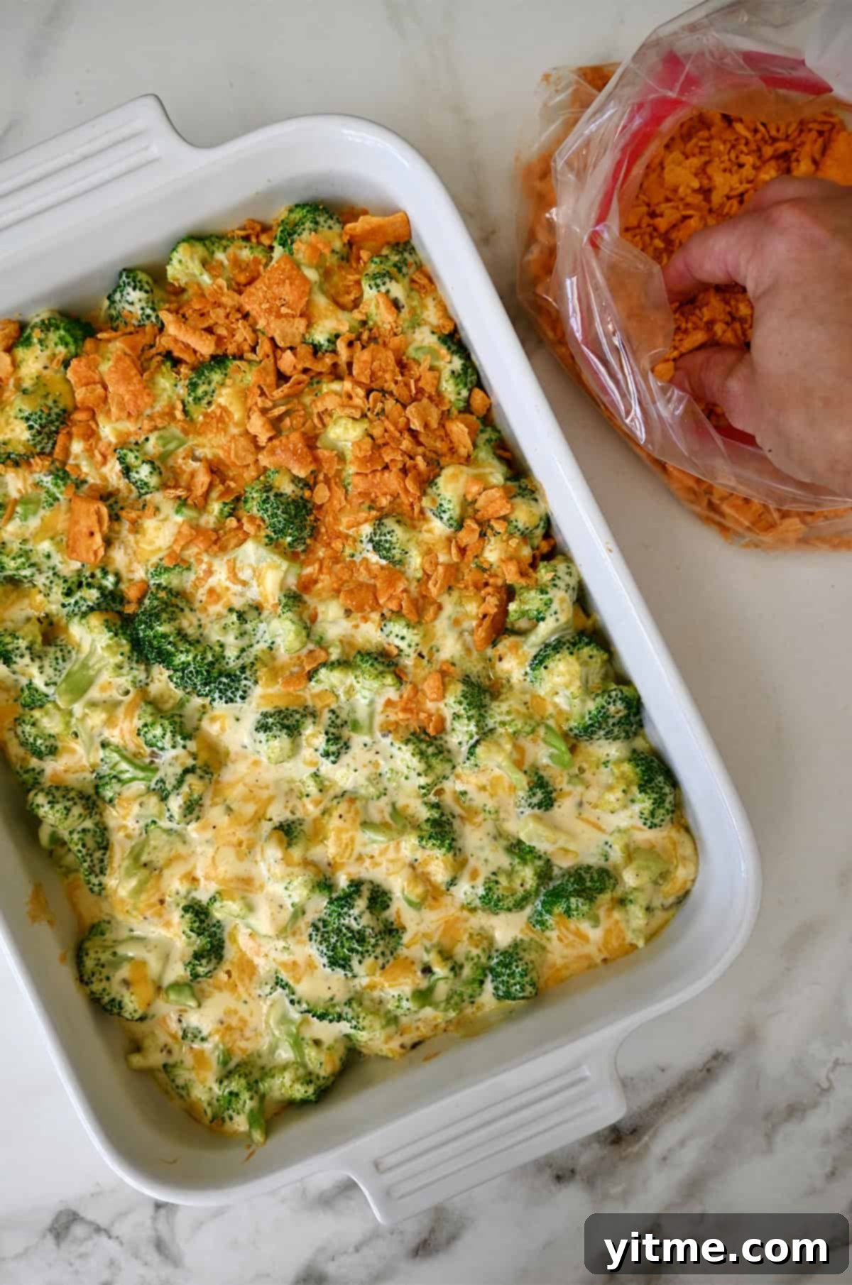 Baked broccoli casserole being topped with crushed cheese crackers. Adding the final touch of crushed crackers.