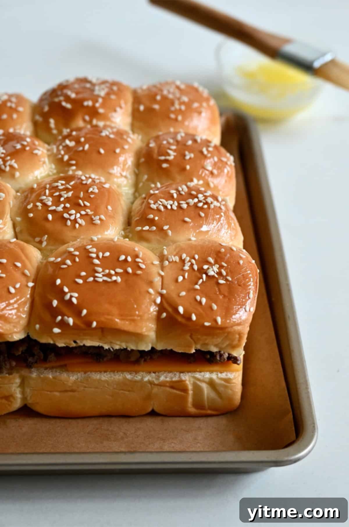 Cheeseburger sliders with sesame seed tops on a parchment paper-lined baking sheet, ready for the oven.
