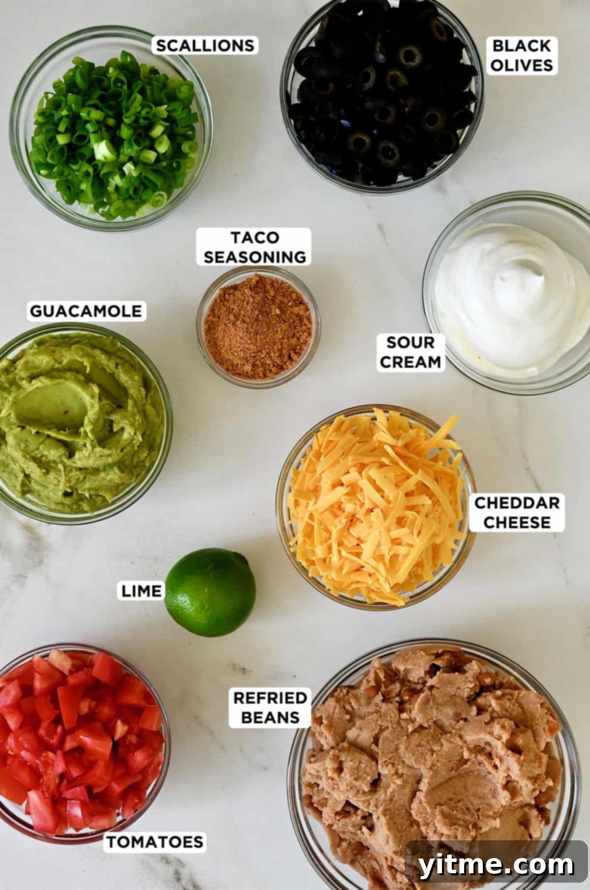 All the colorful ingredients for seven layer dip arranged in glass bowls: refried beans, taco seasoning, sour cream, lime juice, guacamole, diced tomatoes, shredded cheddar cheese, sliced scallions, and black olives.
