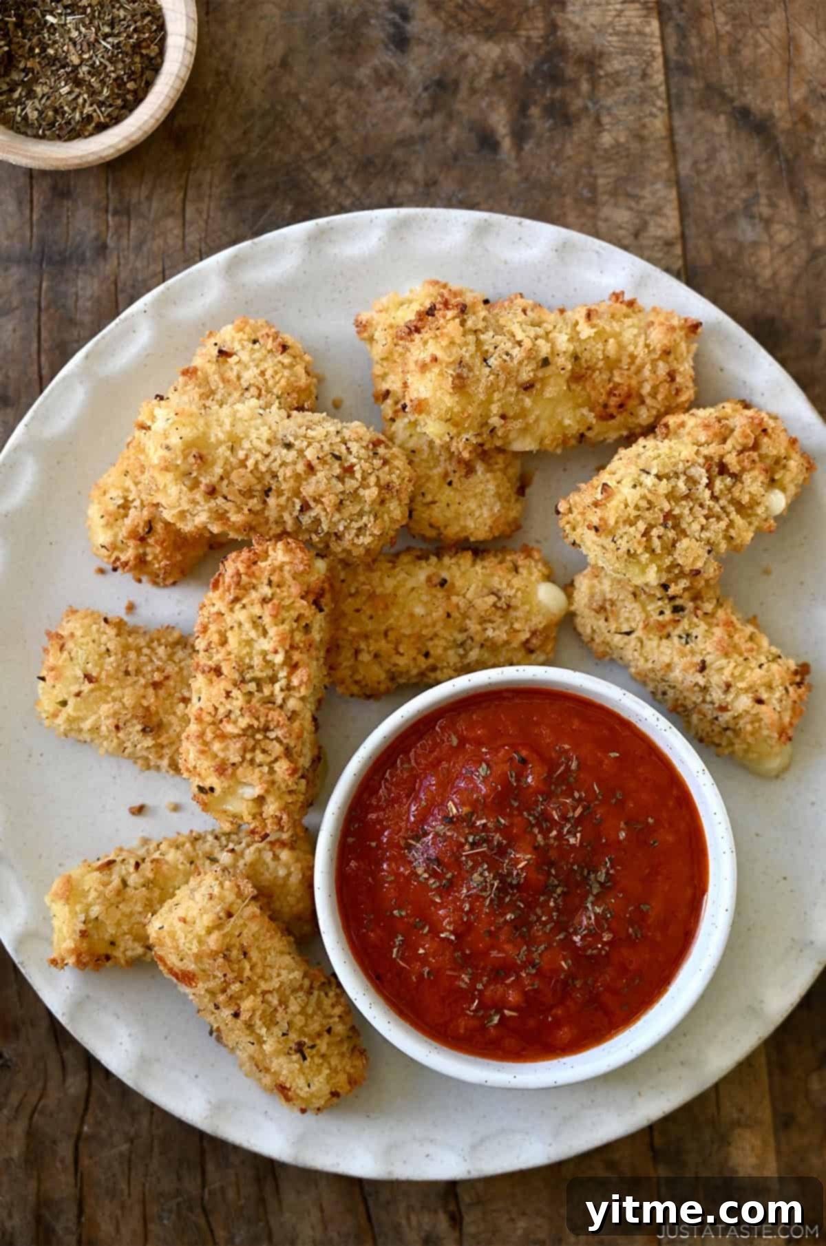 Golden brown mozzarella sticks made in an air fryer on a plate along with a small bowl containing marinara sauce, garnished with fresh parsley.