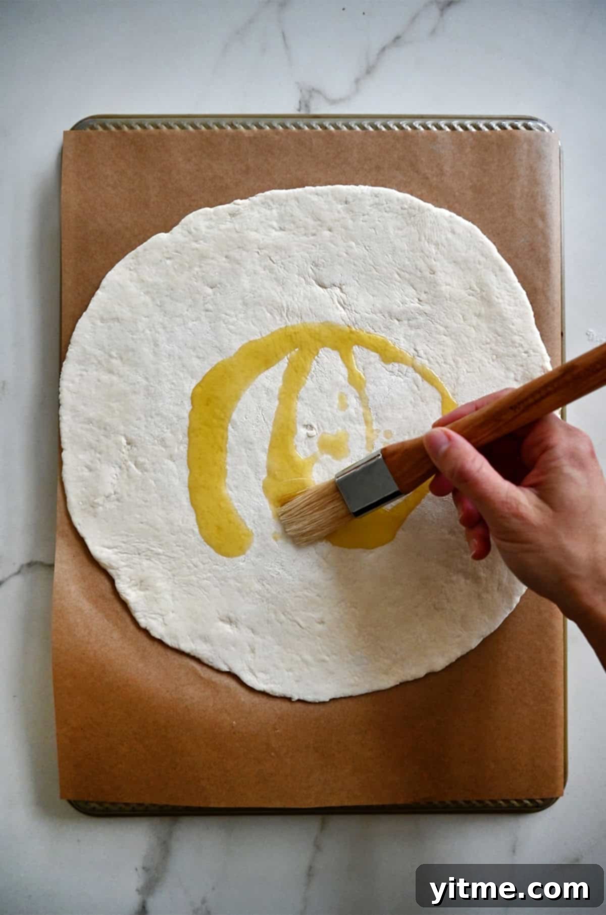 Brushing olive oil atop pizza dough on a parchment paper-lined baking sheet.