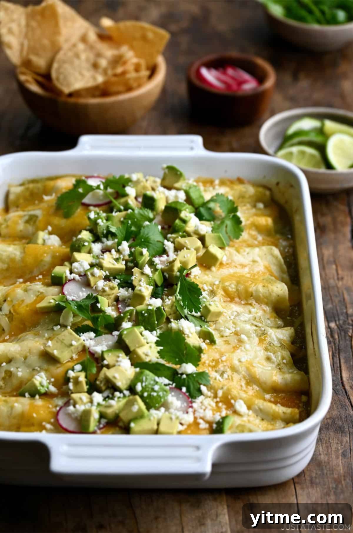Salsa verde enchiladas in a baking dish topped with avocado, radishes, cilantro and cojita cheese. Chips and salsa are nearby.
