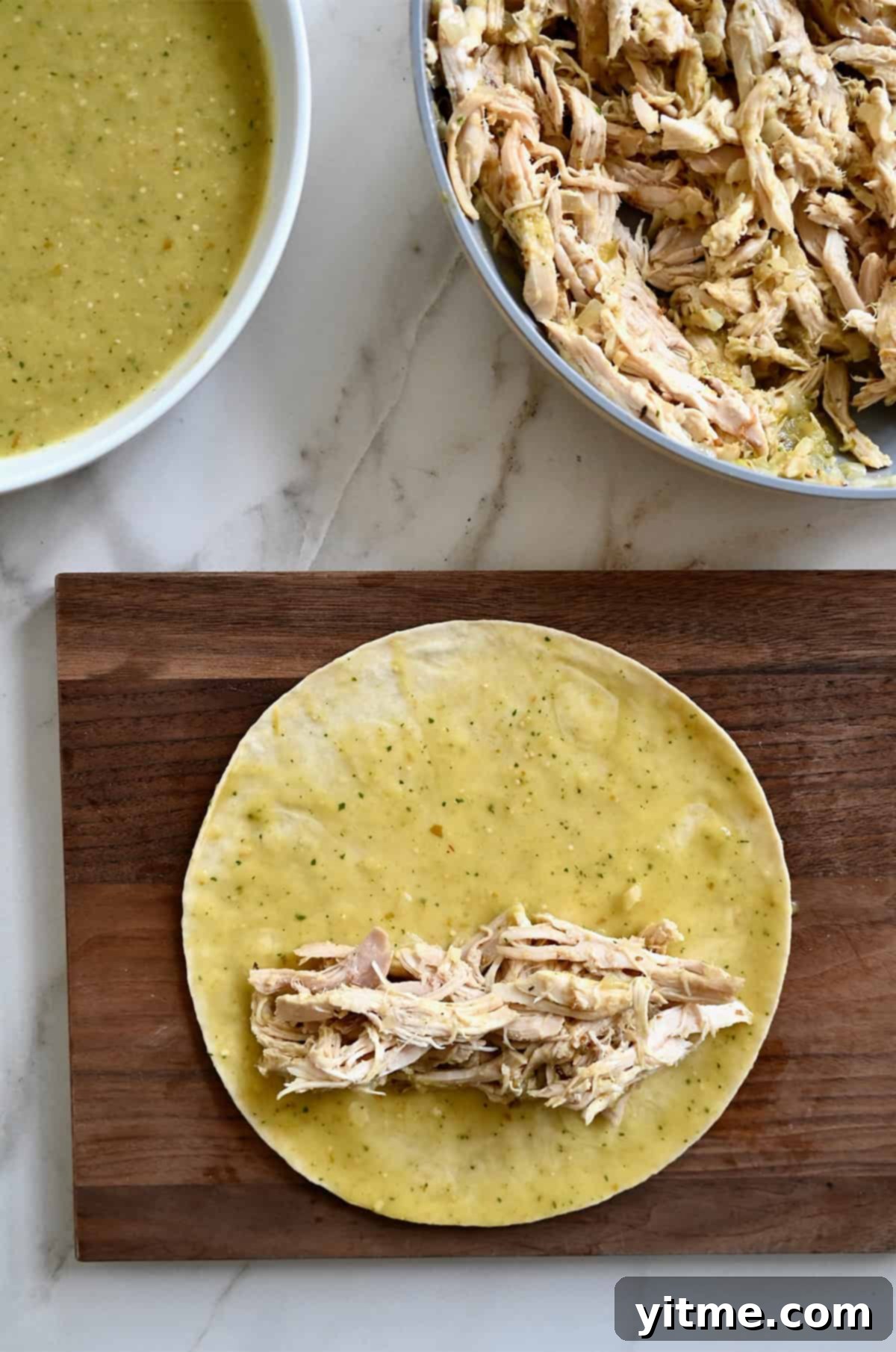 A flour tortilla topped with shredded chicken and green enchilada sauce being assembled on a wooden cutting board, with a bowl of sauce and a pan of chicken nearby.