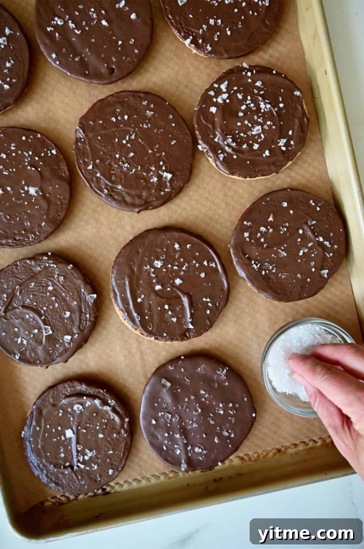 Vanilla Bean Dream Cookies 11 Sprinkling flaky sea salt atop chocolate-dipped cookies on a parchment paper-lined baking sheet. The perfect finishing touch for a balanced sweet and savory experience.
