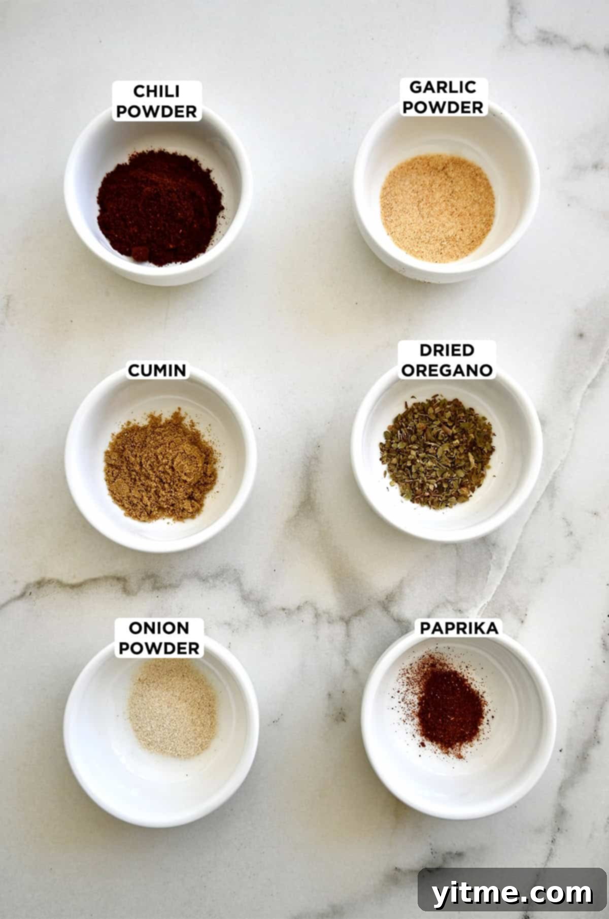 Six small bowls containing taco seasoning ingredients, including chili powder, ground cumin, onion powder, paprika, dried oregano and garlic powder.