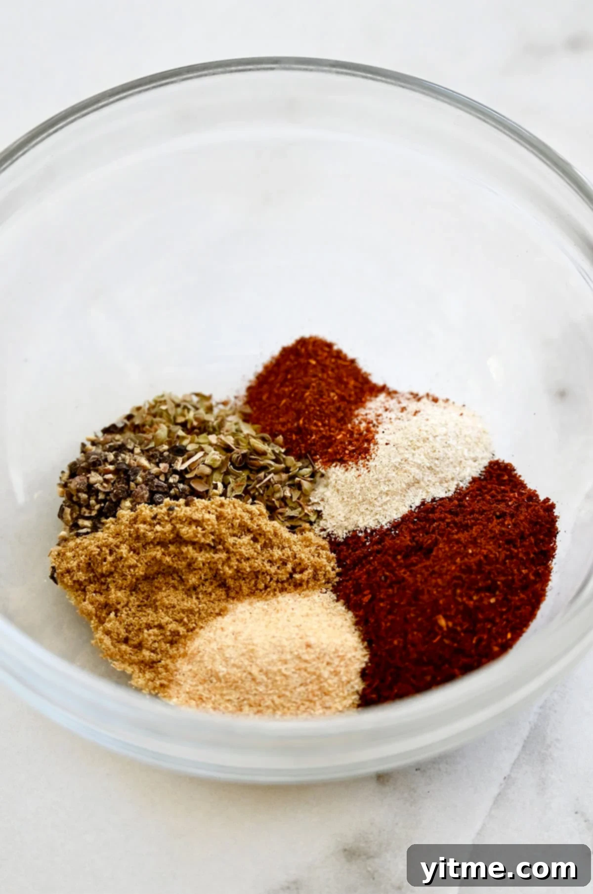 A glass bowl containing garlic powder, garlic onion, chili powder, ground cumin, black pepper, dried oregano and paprika