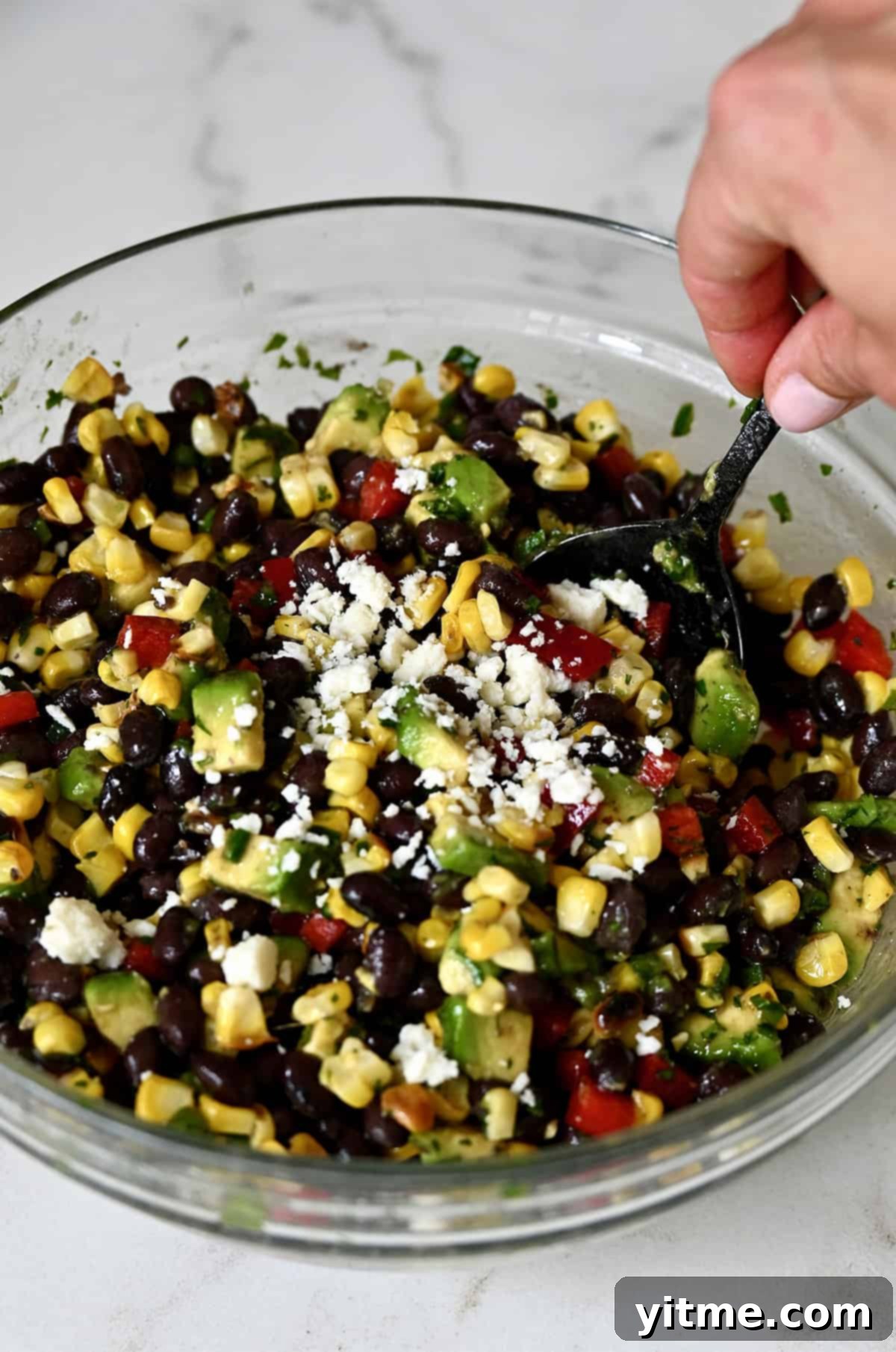A vibrant black bean and corn salad with avocado, tossed in a honey-lime dressing and topped with Cotija cheese, in a glass mixing bowl.