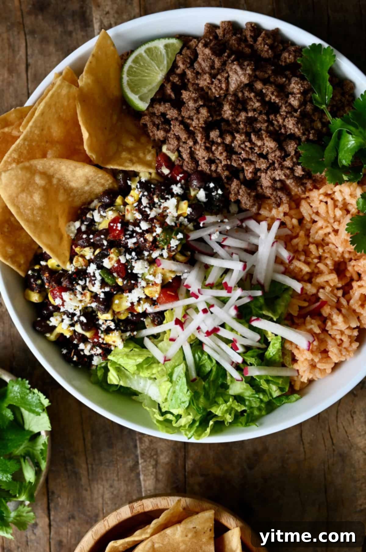 A beautifully assembled taco bowl with shredded lettuce, seasoned ground beef, sliced radishes, black bean and corn salad, fresh cilantro, and crispy tortilla chips.