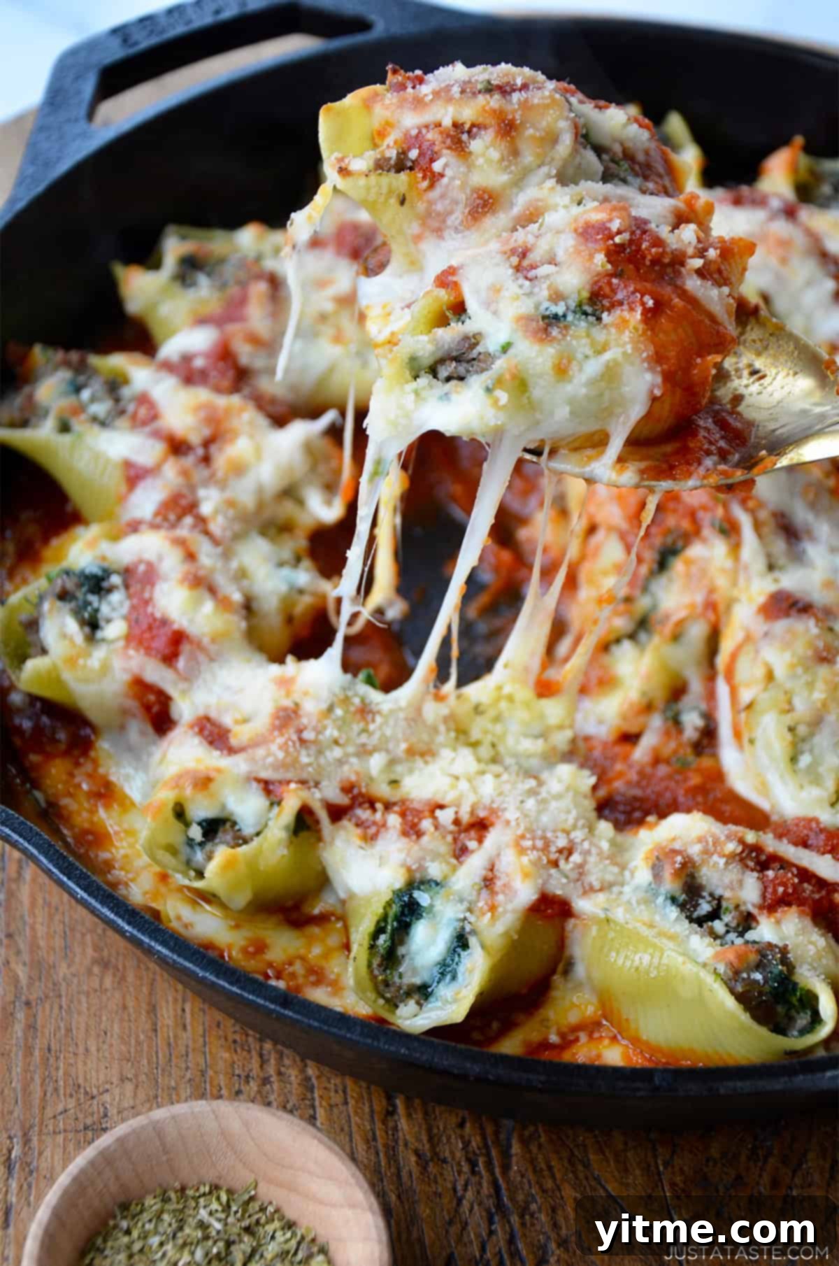 Stuffed shells filled with ground meat, spinach, and ricotta cheese, baked in marinara sauce and topped with melted mozzarella. A serving spoon lifts a shell from a cast iron skillet, revealing gooey, stretchy cheese.