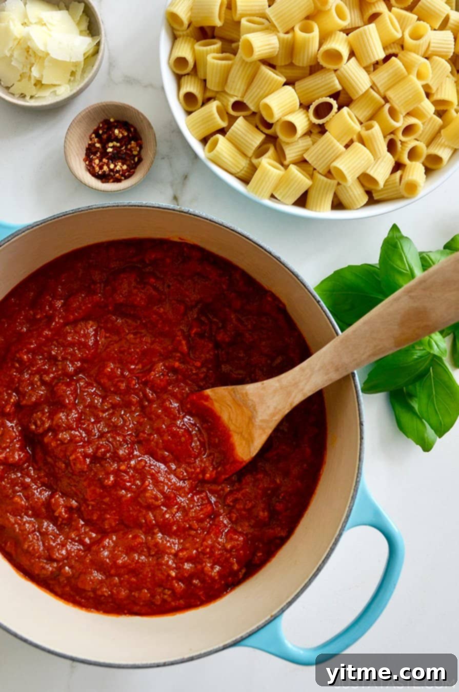 A large stock pot containing Secret Ingredient Bolognese Sauce and a wooden spoon next to a large bowl containing rigatoni noodles