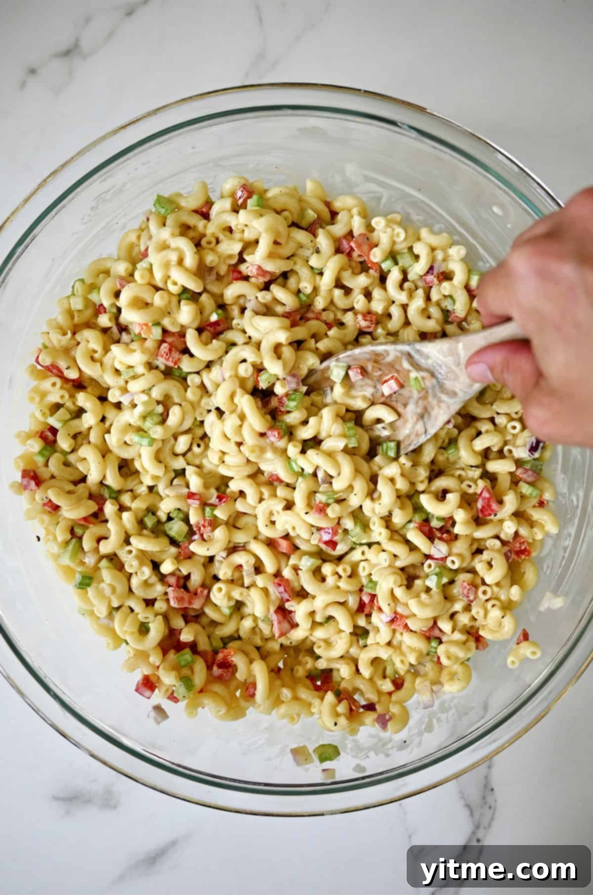 A hand holding a wooden spoon mixes macaroni salad with a creamy dressing in a large bowl.