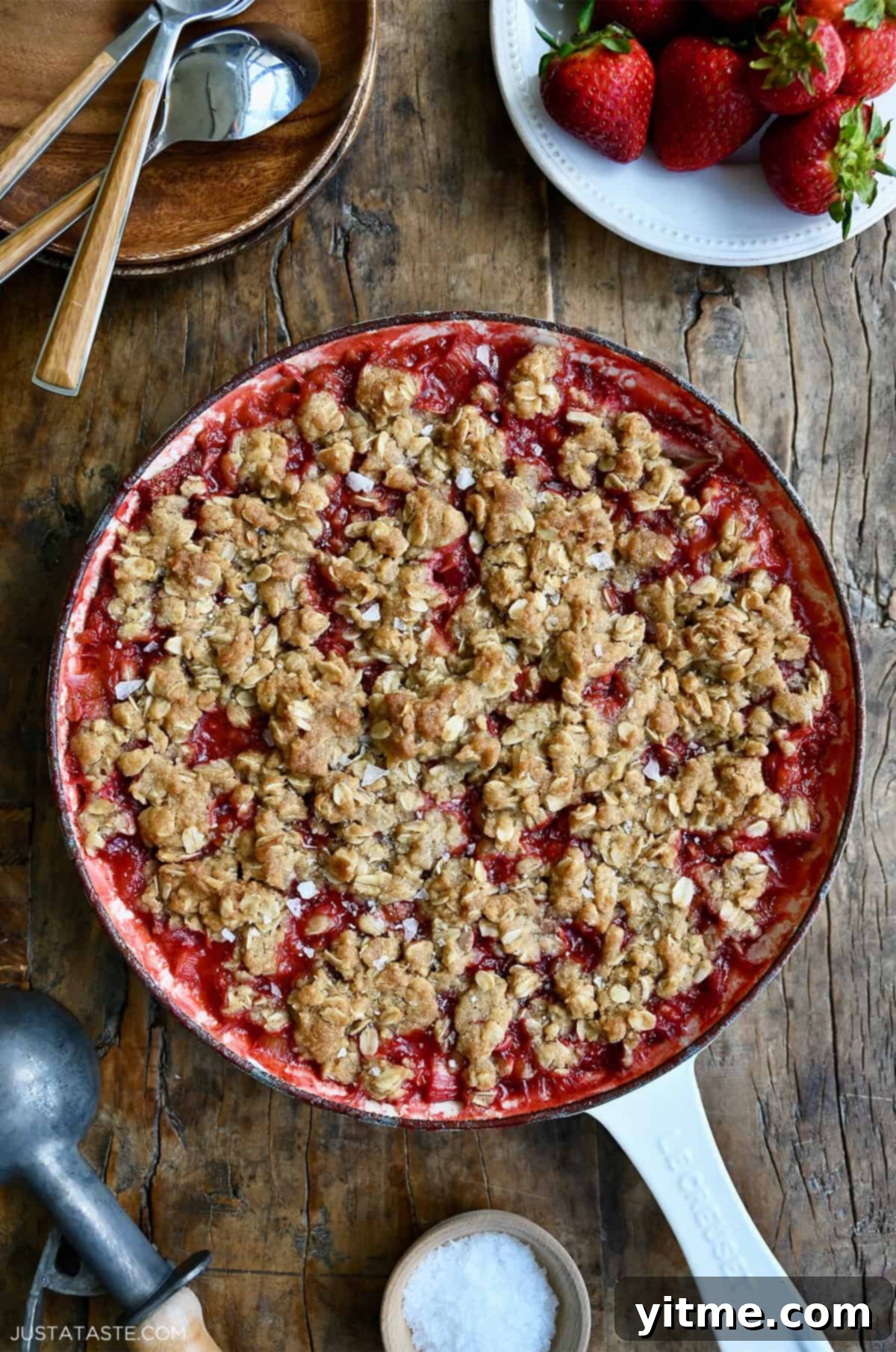 A white enameled cast iron skillet containing strawberry rhubarb crisp. A bowl with fresh strawberries and an ice scoop are nearby - A sweet and tart dessert perfect for summer gatherings.